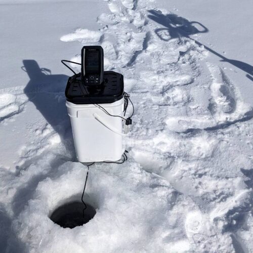 Best ice fishing setup with a plastic bucket containing electronic equipment beside a hole in the ice. Footprints and a snow-covered tool are also visible, enhanced by one of the best ice fishing fish finders.