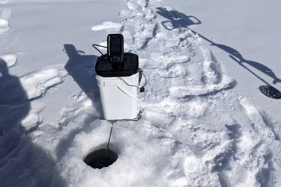 Best ice fishing setup with a plastic bucket containing electronic equipment beside a hole in the ice. Footprints and a snow-covered tool are also visible, enhanced by one of the best ice fishing fish finders.