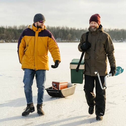 Two people walk on a frozen lake in Michigan, wearing winter clothing. One pulls a sled carrying supplies, including a cooler and an ice auger for ice fishing. Snow-covered landscape and trees are visible in the background.