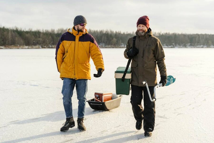 Two people walk on a frozen lake in Michigan, wearing winter clothing. One pulls a sled carrying supplies, including a cooler and an ice auger for ice fishing. Snow-covered landscape and trees are visible in the background.