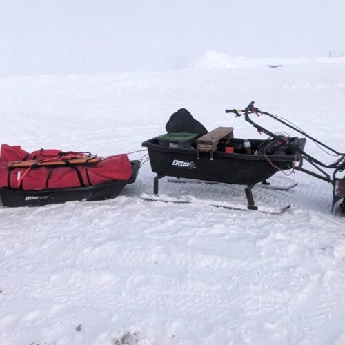 A person dressed in winter gear stands next to a small snowmobile towing three sleds on a snowy landscape, perhaps preparing for ice fishing. Other vehicles are barely visible in the background, hinting at a bustling winter day around the lakes of New York State.