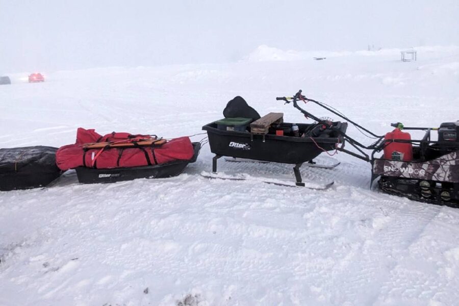 A person dressed in winter gear stands next to a small snowmobile towing three sleds on a snowy landscape, perhaps preparing for ice fishing. Other vehicles are barely visible in the background, hinting at a bustling winter day around the lakes of New York State.