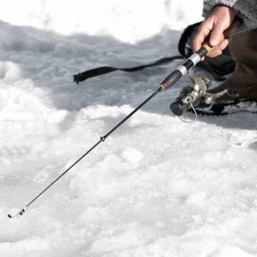 A person holds an ice fishing rod of moderate length while ice fishing on a snowy surface, targeting different sized fish, and wearing brown boots and winter clothing.