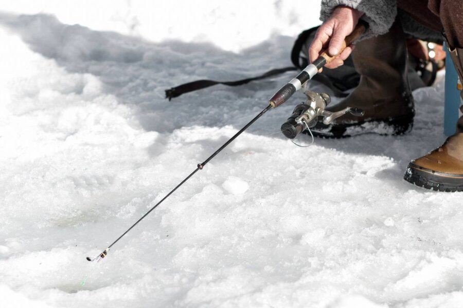 A person holds an ice fishing rod of moderate length while ice fishing on a snowy surface, targeting different sized fish, and wearing brown boots and winter clothing.