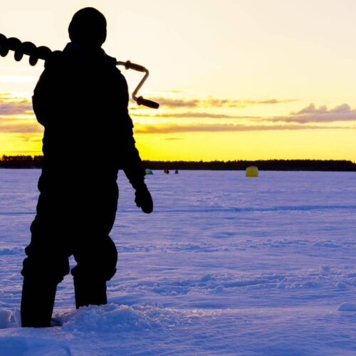 A person stands in the snow holding an ice auger during sunset on a frozen lake, ready for some ice fishing.