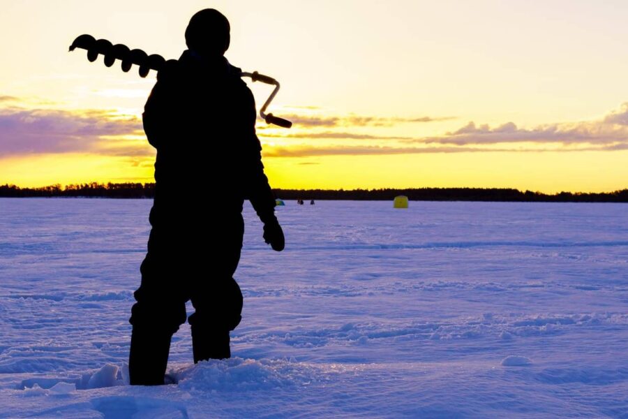 A person stands in the snow holding an ice auger during sunset on a frozen lake, ready for some ice fishing.