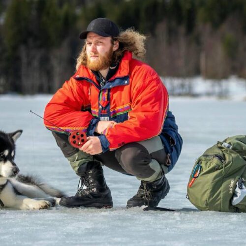 A person in a red jacket and black cap, wearing ice fishing pants, kneels on a frozen lake next to a husky and a large green backpack. Snowy trees and buildings are visible in the background, highlighting the comfort of ice fishing amidst picturesque winter scenery.