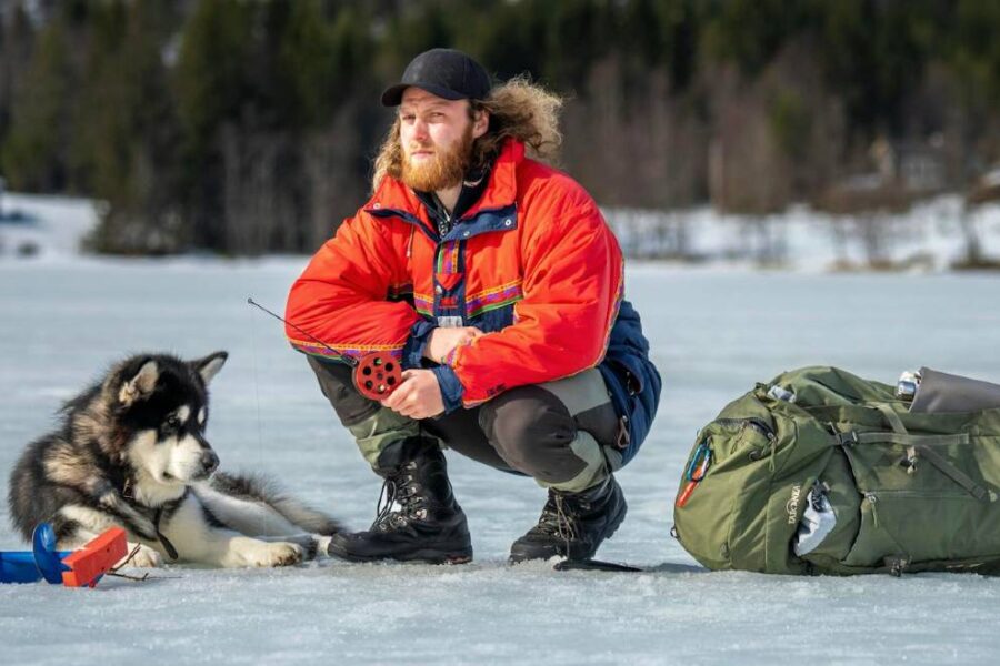 A person in a red jacket and black cap, wearing ice fishing pants, kneels on a frozen lake next to a husky and a large green backpack. Snowy trees and buildings are visible in the background, highlighting the comfort of ice fishing amidst picturesque winter scenery.