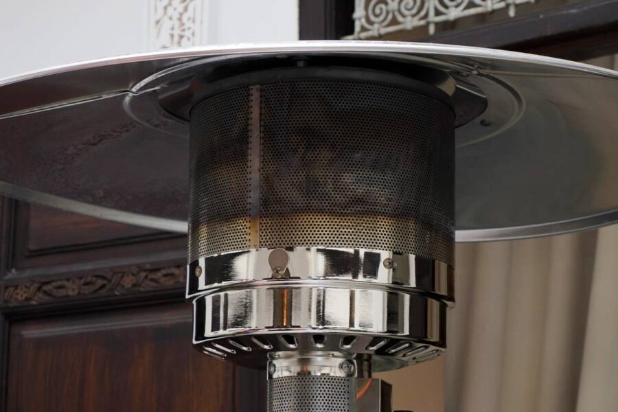 Close-up view of an outdoor patio heater with a metal dome and mesh covering, enhanced by strategic patio heater accessories, positioned in front of a wooden door and draped window.