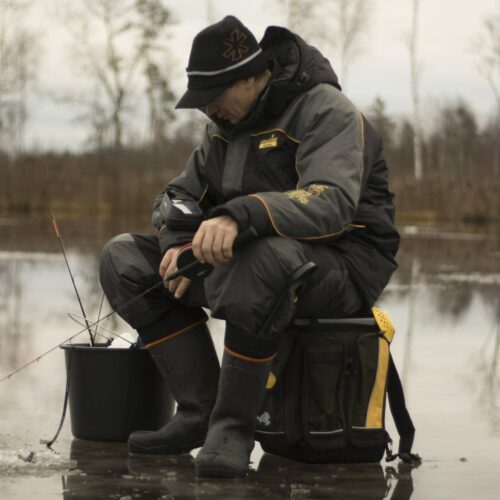 A person dressed in winter gear sits on a bucket on a frozen lake in Pennsylvania with fishing gear, concentrating on an ice fishing hole. Trees are visible in the background, making it one of the best ice fishing lakes around.