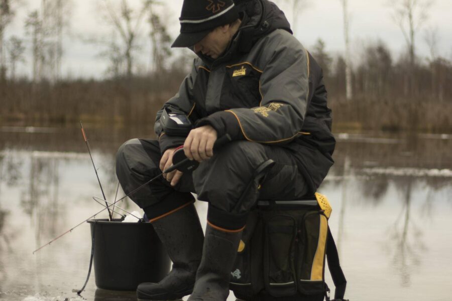 A person dressed in winter gear sits on a bucket on a frozen lake in Pennsylvania with fishing gear, concentrating on an ice fishing hole. Trees are visible in the background, making it one of the best ice fishing lakes around.