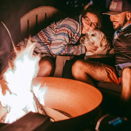 Two people and their furry friend sit around a lit fire pit at night. One person, wearing glasses, hugs the dog, ensuring pet fire pit safety. The other person smiles while wearing a cap.