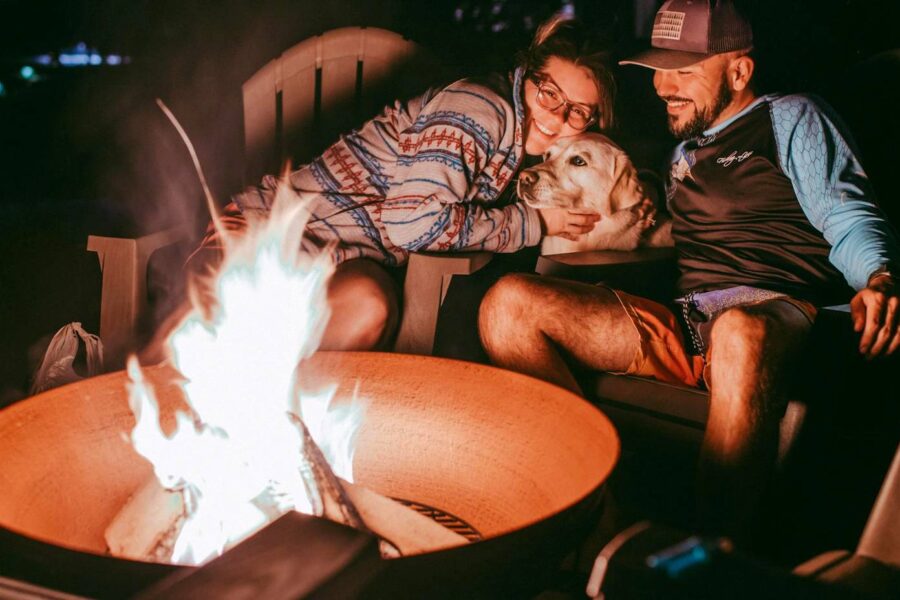 Two people and their furry friend sit around a lit fire pit at night. One person, wearing glasses, hugs the dog, ensuring pet fire pit safety. The other person smiles while wearing a cap.