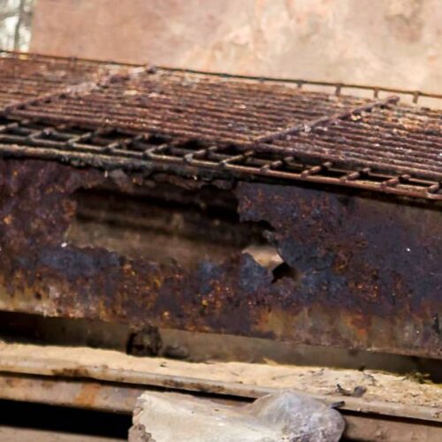 A rusty, rectangular metal charcoal grill with a wire mesh grate sits on a concrete surface, showing visible signs of wear and decay. To prevent this, follow essential prevention tips or remove rust regularly to prolong your grill's life.