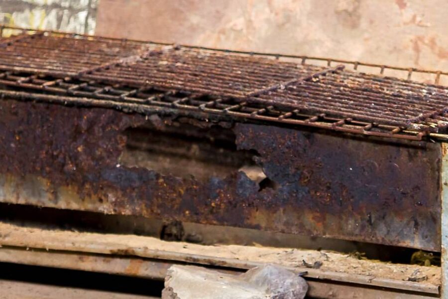 A rusty, rectangular metal charcoal grill with a wire mesh grate sits on a concrete surface, showing visible signs of wear and decay. To prevent this, follow essential prevention tips or remove rust regularly to prolong your grill's life.