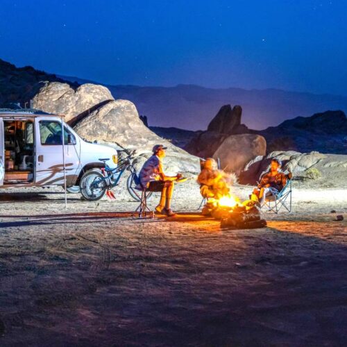 Two camper vans parked at a rocky campsite at night. Four adventurers sit in camp chairs around portable fire pits, with mountains and a starry sky in the background.