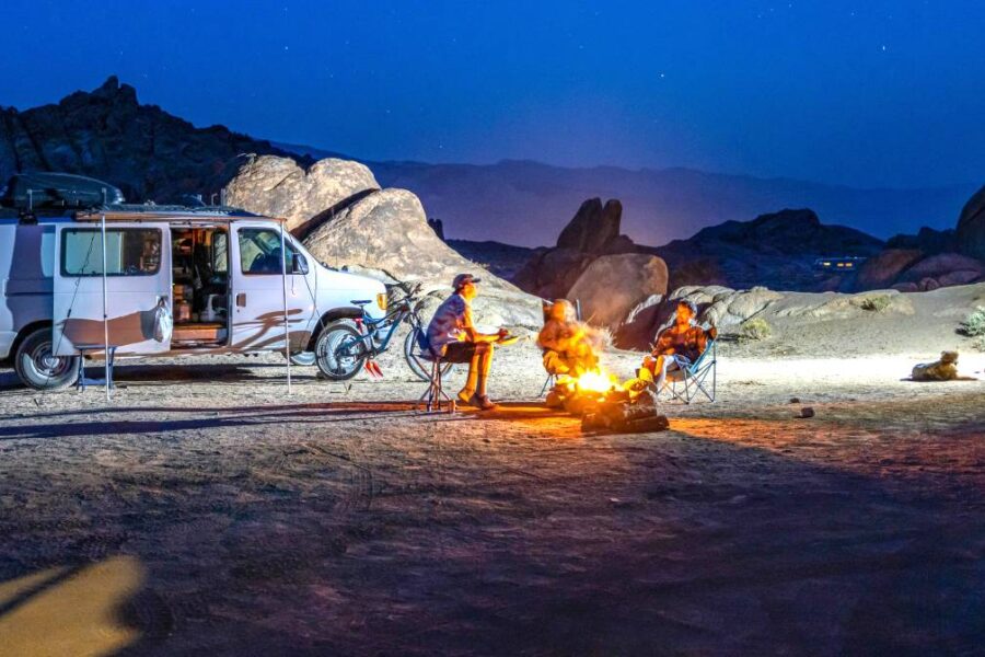 Two camper vans parked at a rocky campsite at night. Four adventurers sit in camp chairs around portable fire pits, with mountains and a starry sky in the background.