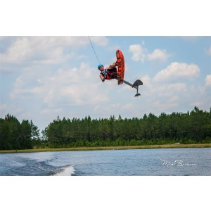 A person wakeboarding and performing an aerial trick above a body of water on a Sky Ski RS Aero 41 Sit-Down Hydrofoil, with a forested background and a partly cloudy sky.