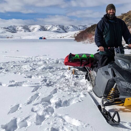 A person stands beside one of the best snowmobiles on a snow-covered landscape with hills and a tent in the background, ready for a day of ice fishing.