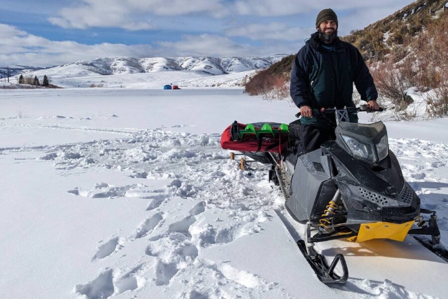 A person stands beside one of the best snowmobiles on a snow-covered landscape with hills and a tent in the background, ready for a day of ice fishing.