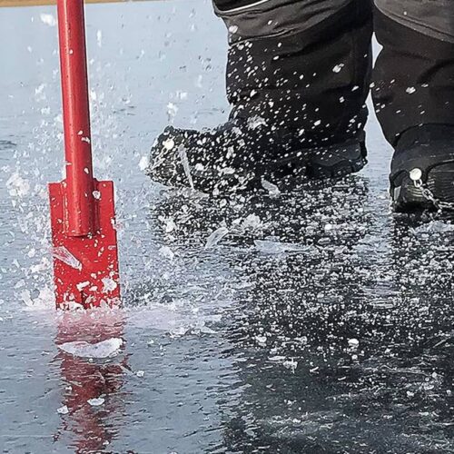 Close-up of a person wearing ice cleats using a red ice auger to drill a hole in the surface of a frozen lake, with an Ice Fishing Spud Bar lying nearby, causing ice shards to scatter.