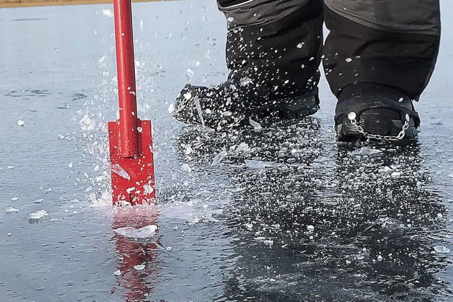 Close-up of a person wearing ice cleats using a red ice auger to drill a hole in the surface of a frozen lake, with an Ice Fishing Spud Bar lying nearby, causing ice shards to scatter.