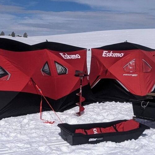 Two red and black Eskimo ice fishing shelters set up on a snowy landscape. Equipment, including sleds, a power auger, and a shovel—essentials for any ice fishing trip—are positioned around the shelters. The sky is partly cloudy.