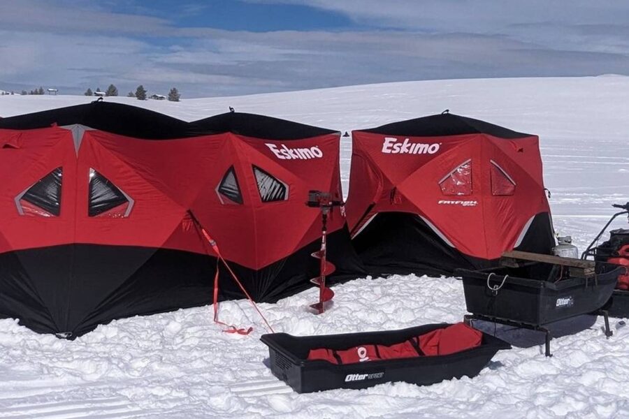 Two red and black Eskimo ice fishing shelters set up on a snowy landscape. Equipment, including sleds, a power auger, and a shovel—essentials for any ice fishing trip—are positioned around the shelters. The sky is partly cloudy.