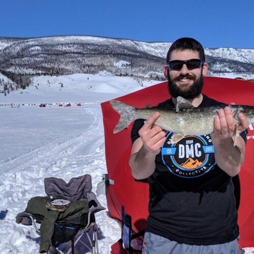 A man stands on a snowy landscape holding a fish with both hands. He is in front of an insulated ice fishing tent, wearing sunglasses and a black t-shirt. Snow-covered mountains are visible in the background, highlighting the frigid weather.