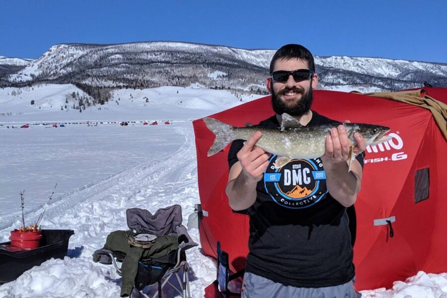 A man stands on a snowy landscape holding a fish with both hands. He is in front of an insulated ice fishing tent, wearing sunglasses and a black t-shirt. Snow-covered mountains are visible in the background, highlighting the frigid weather.