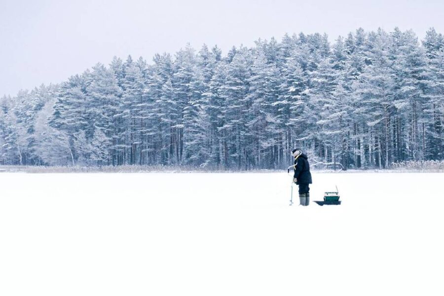 A person stands on a snow-covered field in front of a forest, holding a rod beside a small ice fishing sled, experiencing one of the best ice fishing adventures on the lakes in Idaho.