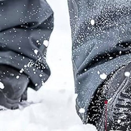 Close-up of a person walking through snow, with visible snowflakes around and snow-covered ice fishing boots. The pants and boots, known for their tread and warmth, appear designed for winter conditions.