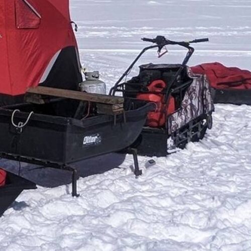 A snowy scene with red tents and equipment set up for ice fishing, including black sleds, a snowmobile, an auger, and utility boxes.