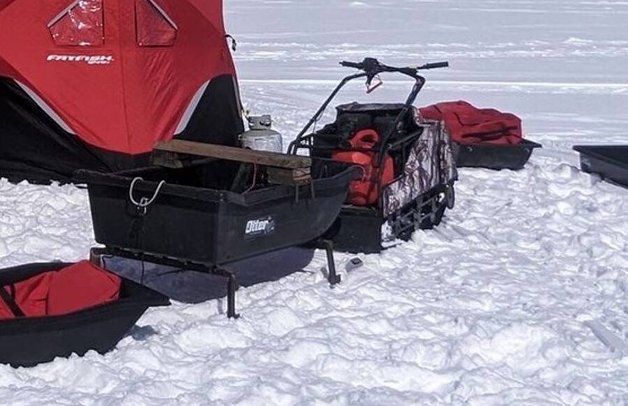 A snowy scene with red tents and equipment set up for ice fishing, including black sleds, a snowmobile, an auger, and utility boxes.