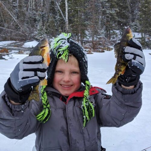 A child in winter clothes holds two fish while ice fishing on the serene lakes of Utah, surrounded by a snowy, wooded area.