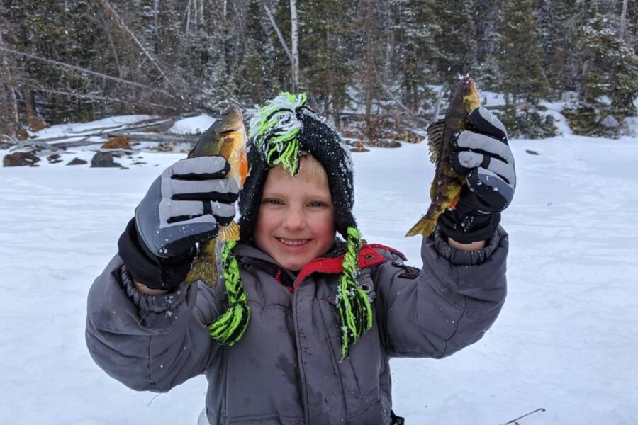 A child in winter clothes holds two fish while ice fishing on the serene lakes of Utah, surrounded by a snowy, wooded area.