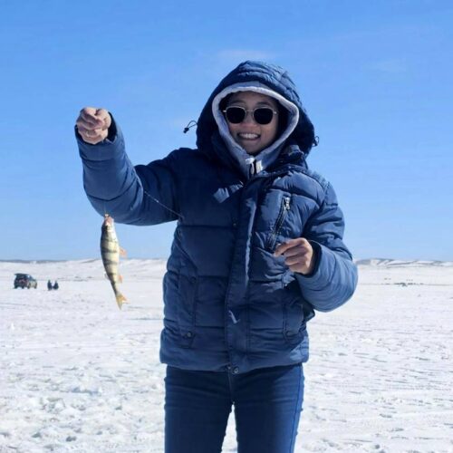 Person wearing a blue jacket, sunglasses, and hood holding up a fish while standing on a snowy landscape with a clear blue sky. Ice fishing equipment and more people are visible in the background, enjoying one of the best ice fishing experiences at lakes in Washington.