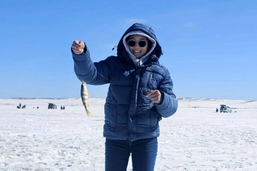 Person wearing a blue jacket, sunglasses, and hood holding up a fish while standing on a snowy landscape with a clear blue sky. Ice fishing equipment and more people are visible in the background, enjoying one of the best ice fishing experiences at lakes in Washington.